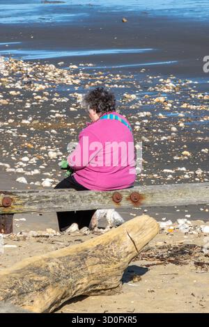 Ältere oder ältere Dame oder Frau, die allein auf einem Holzdampfer an einem leeren Kieselstrand auf der isle of wight in Großbritannien sitzt und sich ausruhen lässt. Stockfoto