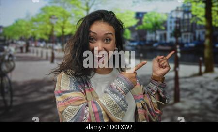 Frau zeigt beide Zeigefinger nach rechts auf einer Straße an einem Kanal und einem baumgesäumten Spaziergang; Freude Verspieltheit. Stockfoto