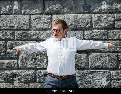 Kaukasischer männlicher Erwachsener in weißem Hemd, der mit ausgestreckten Armen an einer Steinmauer steht. Stockfoto