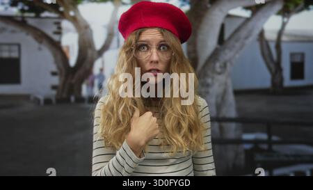Eine Frau, die ihr Haar zeigt, während sie in eine geschäftige Straße zeigt, erscheint als blondes junges Motiv, das eine rote Baskenmütze und eine runde Brille trägt und überrascht aussieht Stockfoto