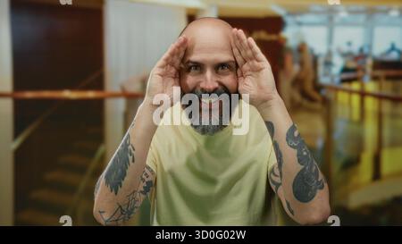 Bärtiger Mann mit Tätowierungen, der das Gesicht in einer Hotellobby verspielt bedeckt und das Charisma und die einnehmende Persönlichkeit im Innenbereich zum Ausdruck bringt. Stockfoto