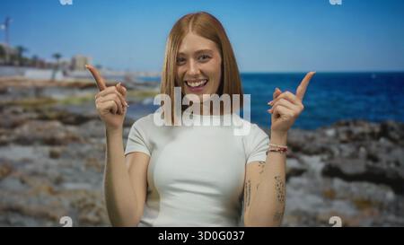 Die junge blonde Frau zeigt die Zeigefinger nach oben auf die felsige Küste an der sonnigen Küste; Optimismus Begeisterung Aufregung Freude. Stockfoto