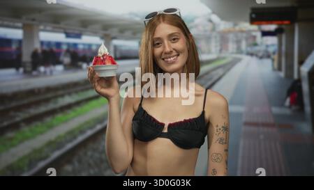 Frau genießt Eis am Bahnhof, trägt Bikini mit Tattoos im Sommer im Freien und hält Dessert bei sonnigem Wetter mit verschwommener Bahn Stockfoto