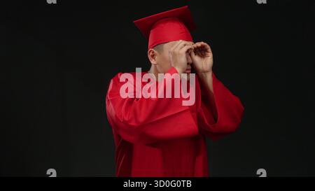 Junger chinesischer Mann in roter Graduierungsmütze und rotem Kleid macht binokulare Gesten isoliert auf schwarzem Hintergrund, die Neugier und Zukunftsvision symbolisieren. Stockfoto
