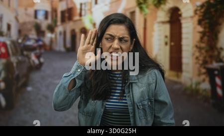 Frau, die ihr Ohr mit der Hand auf einer schmalen Kopfsteinpflasterstraße umhüllt, zuknirscht und einem lauten Geräusch zuzwinkert; Unwohlsein. Stockfoto