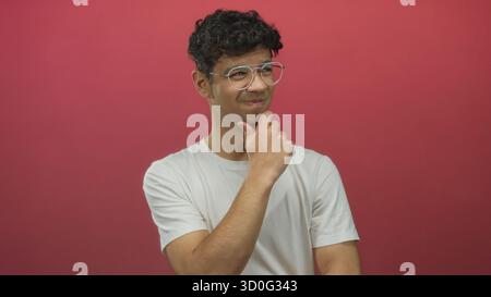 Junger hispanischer Mann mit weißem T-Shirt und runder Brille berührt das Kinn mit nachdenklichem Ausdruck an der roten Studiowand; Zweifel. Stockfoto