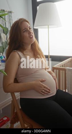 Junge kaukasische Frau, die auf Holzstuhl sitzt und schwangeren Bauch hält und im Kinderzimmer grimmt; Unwohlsein. Stockfoto