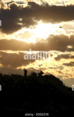 Silhouette einer Person auf einem Hügel mit majestätischem Sonnenuntergang und einem Flugzeug, das über den Hügel fliegt Stockfoto