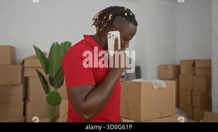 Mann im roten Polo hält Telefon zu Ohr im Haus, umgeben von verpackten Umzugskästen und Pflanzen; bewegender Stress. Stockfoto