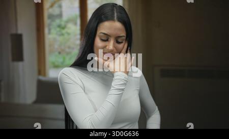 Frau in weiß drinnen, die nachdenklich oder besinnlich wirkt, mit Hand auf der Stirn vor einem weichen Hintergrund, was einen introspektiven Moment in sich andeutet Stockfoto