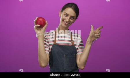 Frau, die roten Apfel in Handschuhen hält und in lila Studio zur Seite zeigt, mit gestreiftem Hemd und Denim Schürze; gesunde Ernährung fröhlich. Stockfoto