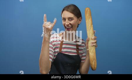 Frau mit Baguette, Schürze und Geste mit Steinhörnern im blauen Studio; verspielte Backenergie. Stockfoto