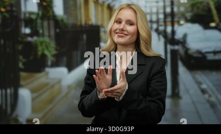 Frau klatscht auf der Straße der Stadt die Hände, zeigt Jugendliche Energie und professionelle Kleidung in urbaner Umgebung mit blondem Haar, was eine selbstbewusste und stilvolle Atmosphäre schafft Stockfoto