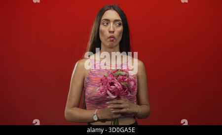 Frau mit rosa Blumenstrauß und gepucktem Fisch im Studio mit rotem Hintergrund; verspielter Neugier-Charme. Stockfoto