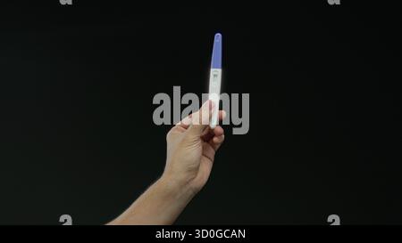 Mann hält Schwangerschaftstest über schwarzem Hintergrund, Hand isoliert, zeigt Ergebnis in hellem Raum, erfasst einen intimen und signifikanten Moment in Heilung Stockfoto