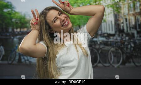 Die sonnenbeschienene Frau lächelt spielerisch und zeigt mit ihren Fingern ein doppeltes Friedenszeichen an einem Kanal unter hellem Tageslicht; Glück. Stockfoto