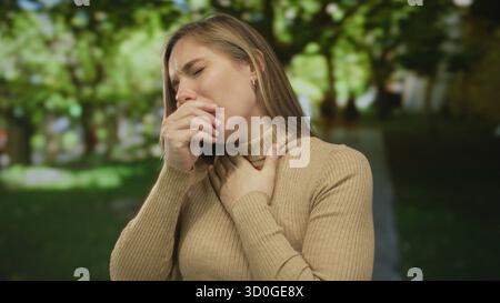 Frau draußen im Park, die Beschwerden oder Schmerzen zeigt, Kehle umklammert, umgeben von Bäumen und Gras an einem sonnigen Tag in lässiger Kleidung. Stockfoto