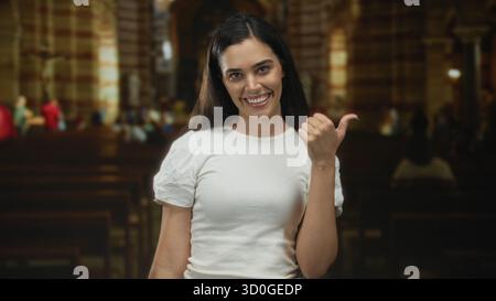 Die hispanische junge Frau zeigt mit Daumen zur Seite im Kirchengebäude, lächelt und steht an Holzbänken unter warmem Innenlicht; einladende Freude. Stockfoto