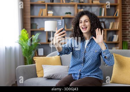 Junge Frau mit lockigen Haaren auf einem Sofa lächelt und winkt während eines fröhlichen Smartphone-Videoanrufs und genießt legere Ferngespräche mit Freunden oder Familie von zu Hause aus Stockfoto