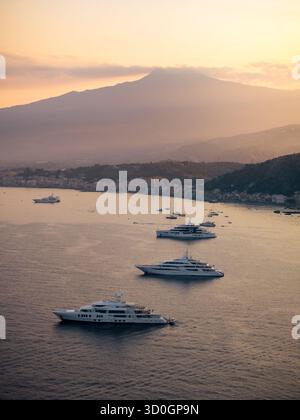 Blick aus der Vogelperspektive auf luxuriöse Yachten, die über das ruhige Wasser vor dem Hintergrund des Ätna und der Küste in der Abenddämmerung gleiten, Taormina, Sizilien, Italien. Stockfoto