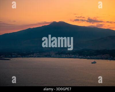 Aus der Vogelperspektive erhebt sich der Ätna majestätisch über der Küste, während die Sonne untergeht und ein warmes orangefarbenes Leuchten über den Himmel wirft, Taormina, Sizilien, Italien. Stockfoto