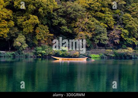 Ein einsames Holzboot ruht sanft auf einem ruhigen, smaragdgrünen Fluss und spiegelt die dichten Baumkronen des umliegenden Waldes wider. Der goldene Farbton des Bootsverbandes Stockfoto