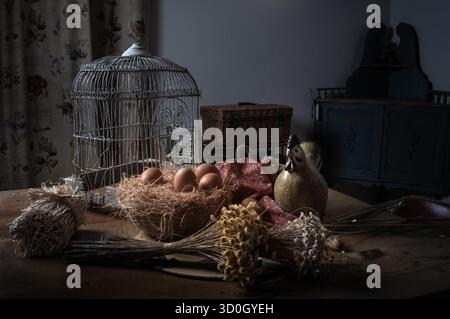 Rustikales Stillleben mit Eiern im Strohnest, getrockneten Blumen, Korb und Vintage-Vogelkäfig auf Holztisch. Landhausatmosphäre und Tradition Stockfoto