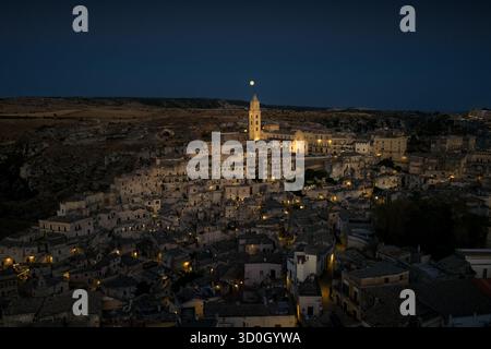 Blick aus der Vogelperspektive auf alte, in goldenes Licht getauchte Wohnungen unter einem ruhigen Nachthimmel, mit dem Turm des Duomos, der zum Mond reicht, Matera, Basilicata, Italien. Stockfoto