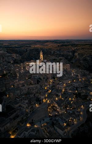 Aus der Vogelperspektive auf alte Steinbauten, die unter einem pastellfarbenen Himmel mit warmem Licht leuchten, erhebt sich der Turm des Duomo in Matera, Basilicata, Italien. Stockfoto