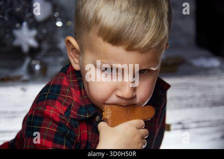 Das entzückende kaukasische Kind isst Lebkuchen und macht einen lustigen Gesichtsausdruck. Weihnachtsessen. Stimmungsvolles, ästhetisches Weihnachtshaus für Silvester. Fröhlicher Junge. Fröhlich, gute Laune. Stockfoto