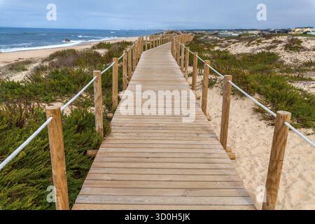 Eine hölzerne Promenade mit Seilgeländern schlängelt sich durch Sanddünen und grüne Sträucher in Richtung eines ruhigen Strandes unter einem bewölkten Himmel Stockfoto