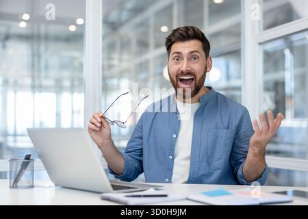 Geschäftsmann im modernen Büro-Laptop reagiert mit freudiger Überraschung und hebt seine Hand, nachdem er gute Nachrichten über Promotion, Deal oder erfolgreiche Investitionen gelesen hat Stockfoto