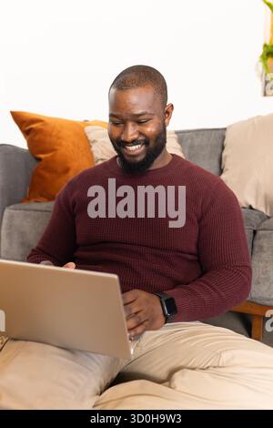 Mittelalter afroamerikanischer Mann, der auf dem Boden sitzt und auf dem Sofa auf silbernem Laptop in der Nähe der Pflanze tippt Stockfoto