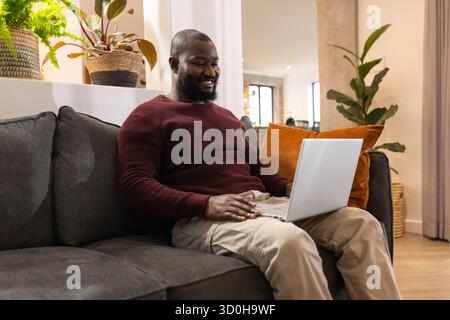 Afroamerikanischer Mann sitzt auf dunkelgrauem Sofa unter Zimmerpflanzen im Wohnzimmer und arbeitet am Laptop Stockfoto