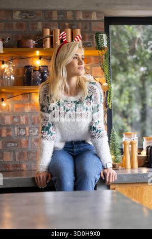 Frau, die auf einer Steinplatte in der gemütlichen Küche sitzt und einen Urlaubspullover und ein Stirnband mit Zuckerrohr trägt Stockfoto