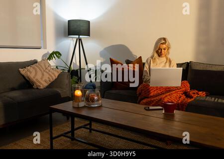 Frau tippt am Laptop und kuschelt sich unter der Decke neben dem Couchtisch im Wohnzimmer, Kopierraum Stockfoto
