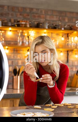 Frau, die herzförmige Kekse mit Glasbeutel auf der Arbeitsplatte in der Wohnküche unter Ringlicht dekoriert Stockfoto