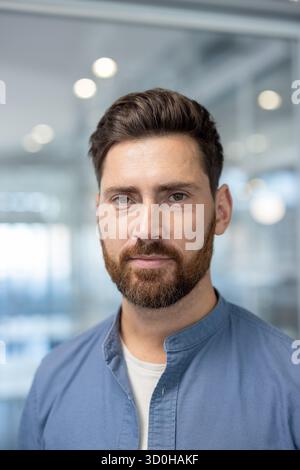 Selbstbewusster bärtiger Geschäftsmann mittleren Alters in einem hellen, modernen Büro, klug-lässige Führungspositionen, die Führungsstärke, Erfolg, Entschlossenheit und professionelles Charisma vermitteln Stockfoto