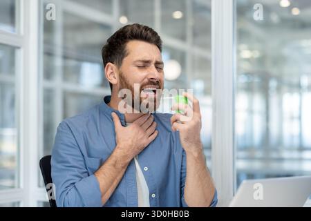 Bärtiger Mann an seinem Schreibtisch, der Rachenmedizin sprüht, um Halsschmerzen und Beschwerden während der Arbeit zu lindern, Nahaufnahme zeigt Selbstversorgung während der Krankheit an einem modernen Arbeitsplatz Stockfoto