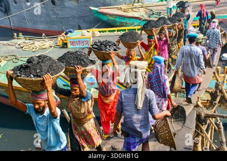 Dhaka, Bangladesch - 15. April 2024: Blick auf Arbeiter, die Körbe mit Kohle auf ihren Köpfen ausbalancieren, über eine enge Holzbrücke in der Nähe von Booten gehen, ein Vibr Stockfoto
