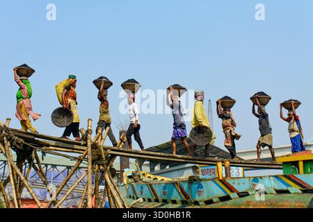Dhaka, Bangladesch - 15. April 2024: Blick auf Arbeiter, die Körbe mit Kohle auf ihren Köpfen ausbalancieren und provisorische Bambusbrücken über lebendigen, bemalten Booten unter klarem Himmel durchqueren. Stockfoto