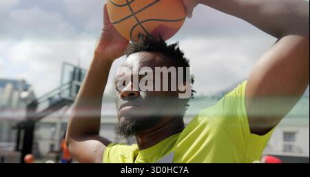Männlicher Athlet in gelbem Hemd, der Basketball über dem Basketballfeld hält, mit Korb, Rückenbrett Stockfoto