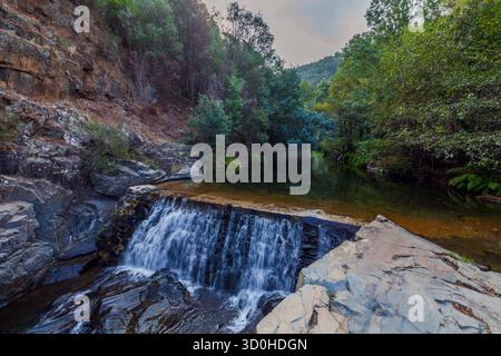 Ein kleiner, kaskadierender Wasserfall stürzt über dunkle Felsen in ein ruhiges Flussbecken, umgeben von üppig grünen Wäldern und einer zerklüfteten Klippe unter einer bewölkten Oberfläche Stockfoto