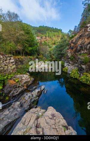 Ein kleiner Wasserfall fällt über dunkle, geschichtete Felsen in einen rauschenden Fluss, der von üppig grünen Wäldern und zerklüfteten Klippen unter einem bewölkten Himmel eingerahmt wird Stockfoto