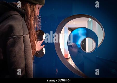 Ein Mädchen in einem dunklen Hoodie und Hut interagiert mit einer blauen Wand mit einem großen spiralförmigen Ausschnitt und kreisförmigen Öffnungen. Stockfoto