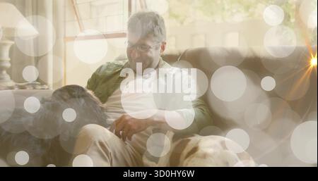 Mann mittleren Alters mit Brille, olivfarbenem Hemd, streichelt zwei Hunde auf Sofa im Wohnzimmer mit Lampe Stockfoto