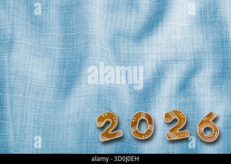 Süße 2026-Nummer für frohe Neujahrsgrüße auf blauem welligem Textilinhalt. Braune Lebkuchenkekse mit weißer Glasur auf wunderschöner Leinwand. Stockfoto