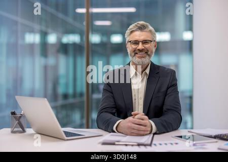 Erfahrener Geschäftsmann mit Brille und Anzug, sitzt selbstbewusst mit Laptop und Dokumenten am Schreibtisch und lächelt in die Kamera in einem modernen Büroumfeld Stockfoto