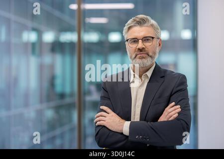 Reifer Geschäftsmann, der mit überkreuzten Armen in einem modernen Firmenbüro steht, ernsthaft in die Kamera schaut und Selbstvertrauen, Professionalität und Erfahrung am Arbeitsplatz vermittelt Stockfoto