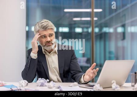 Reifer Geschäftsmann, der Stress und Frustration erlebt, während er an einem Laptop arbeitet, umgeben von zerknitterten Papieren, mit einer herausfordernden Aufgabe oder einem kreativen Block zu kämpfen hat Stockfoto